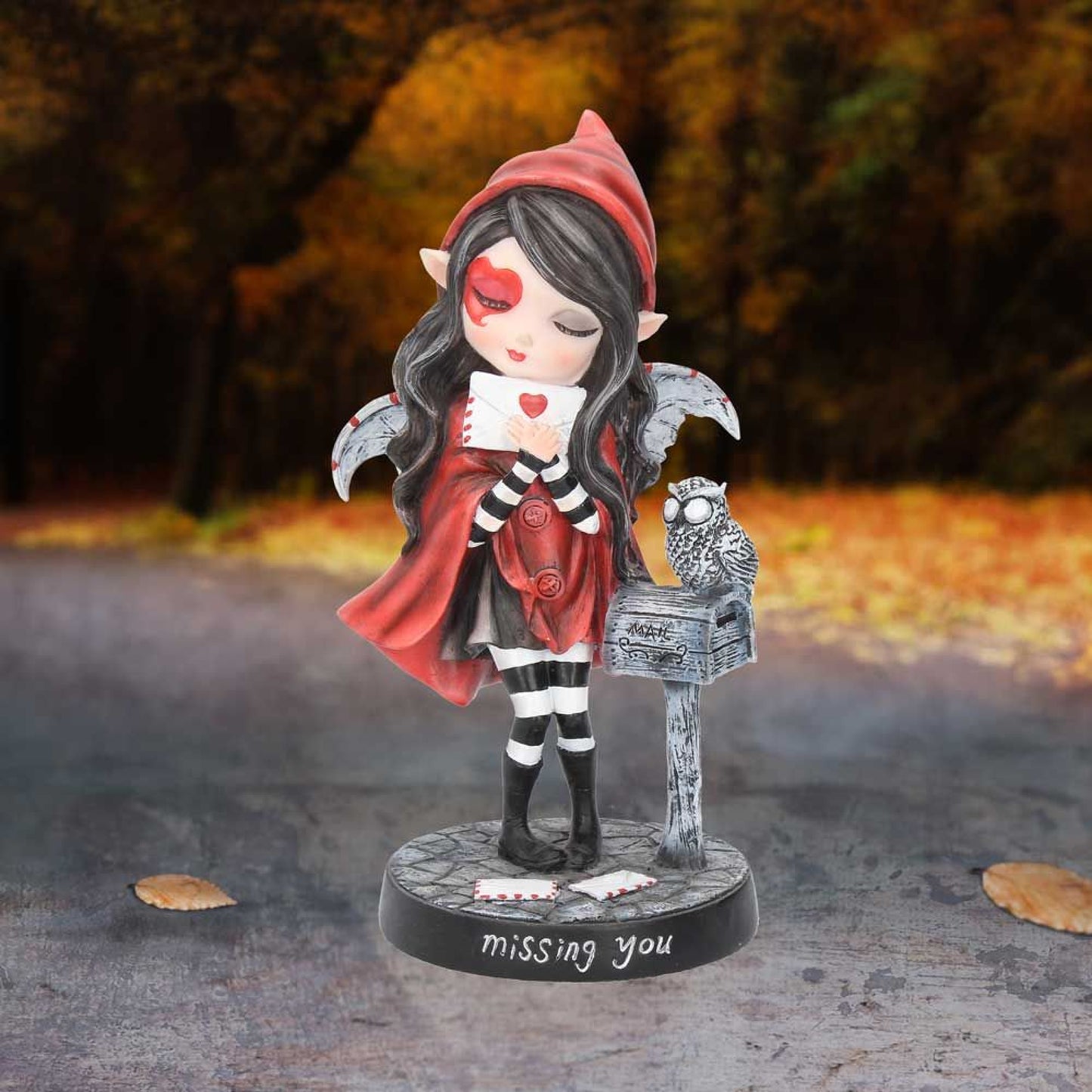Wearing a red, hooded cloak this unique Fairy has a pair of delicate white wings emerging from her back.  Her raven hair frames her face which is decorated by a large heart.  In her hands, she holds a love letter close while her owl companion sits calmly on a wooden letter box.  The theme behind the figurine is emblazoned on the black base, ‘Missing You’. 17.5cm high